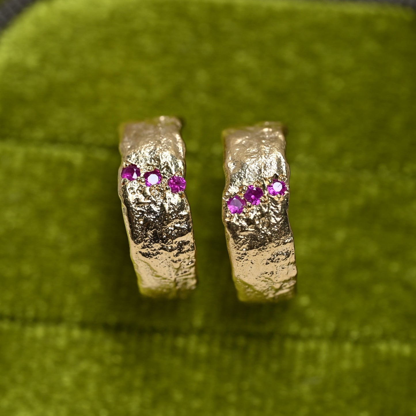 A pair of textured gold hoop earrings with three small pink gemstones on each earring, displayed on a green velvet surface.