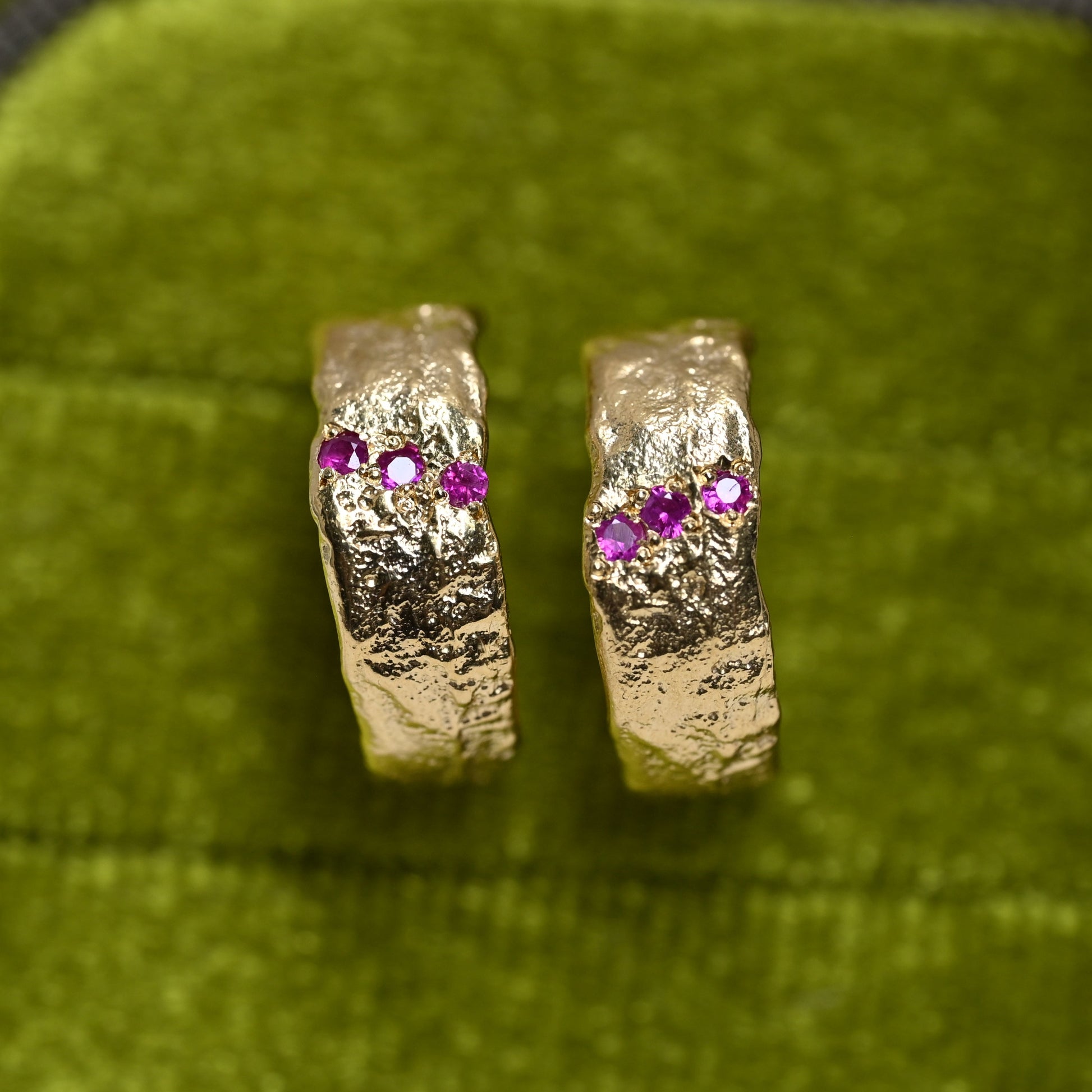 A pair of textured gold hoop earrings with three small pink gemstones on each earring, displayed on a green velvet surface.