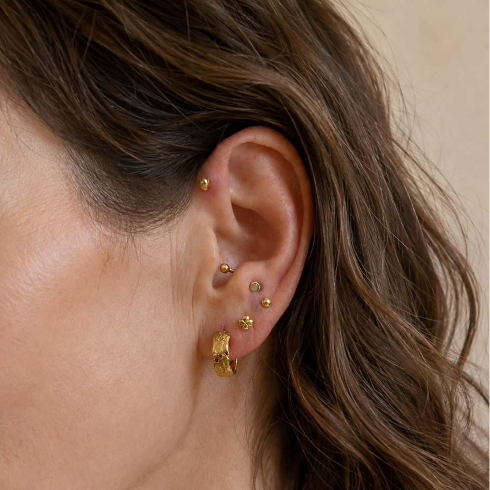 A close-up of a woman's ear wearing multiple gold ear piercings, including studs and a small gold leaf cluster earring, against a soft beige background.
