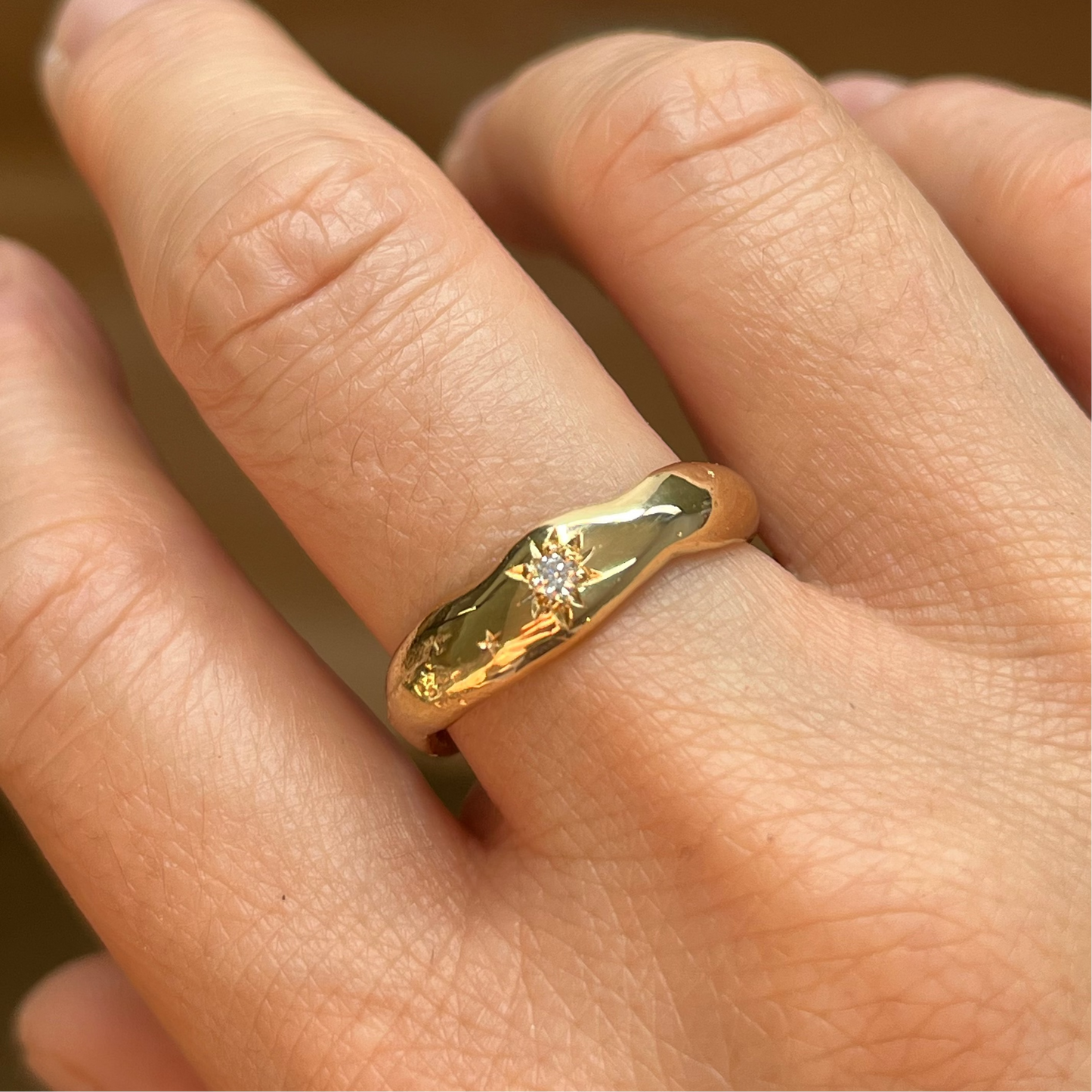 A close-up of a hand wearing a gold ring with a small central clear gemstone and engraved star-like details on the top.