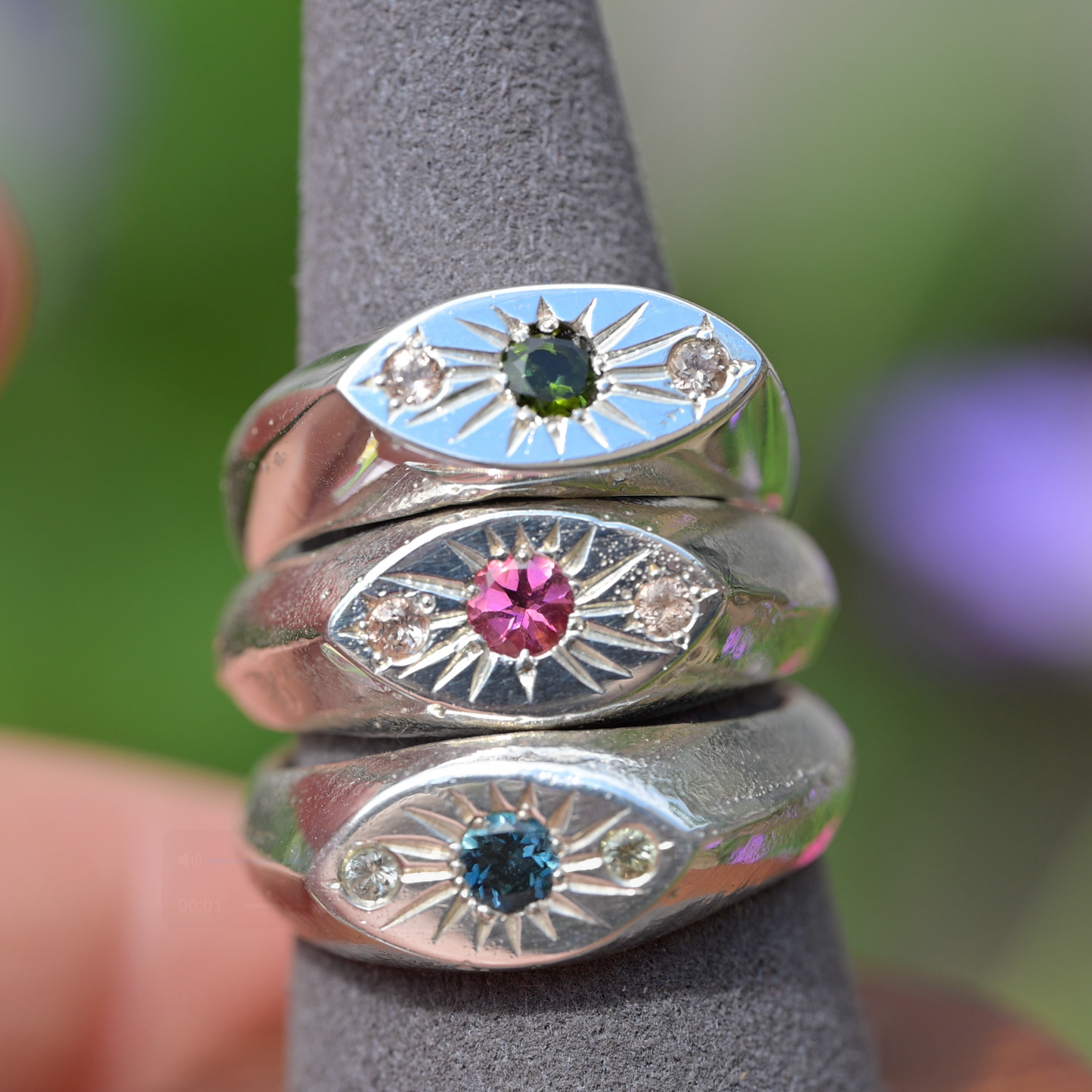 Three silver evil eye rings with different colored gemstones (green, pink, and blue) displayed on a gray ring stand.